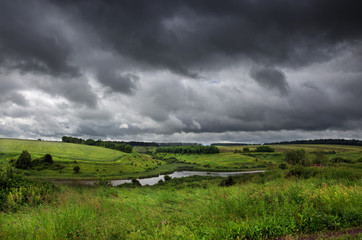 Cloudy summer landscape with small river.Dark stormy clouds in dramatic overcast sky.Fields,green...