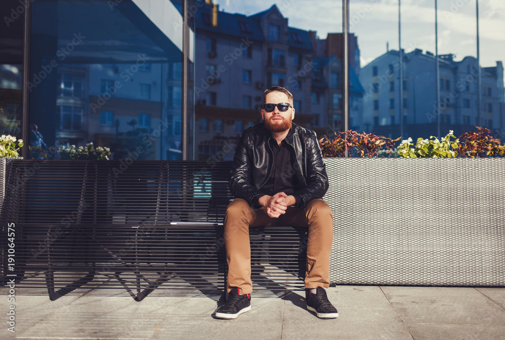 Wall mural Bearded man in a leather jacket and sunglasses sits on a bench