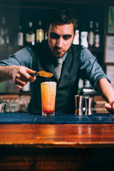 preparation of perfect summer cocktail, orange with cherry and orange slice. Close up of handsome bartender at work