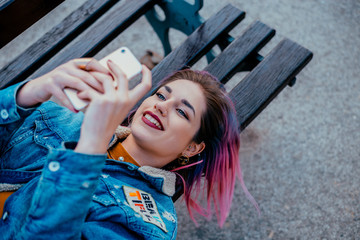 Smiling girl with purple hair lying on a bench, using phone. - obrazy, fototapety, plakaty