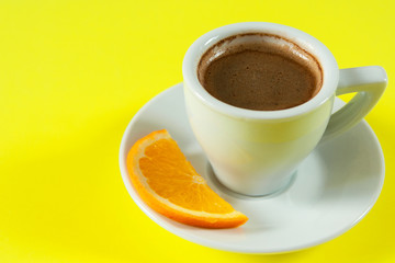 A cup of espresso with foam on a white saucer with an orange slice. Bright yellow background. Close-up. Place for text