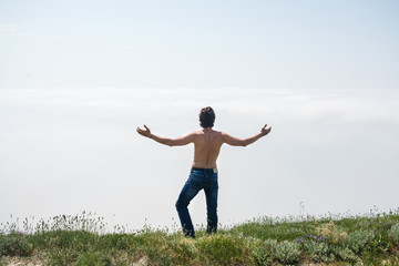 Young man with naked torso in jeans and glasses is standing on the edge of a cliff and looks at the fog.
