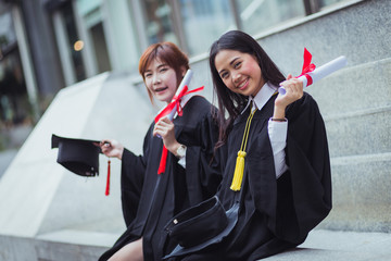 Portrait of Two happy graduated young girls in graduation gowns holding diplomas and smile together in city