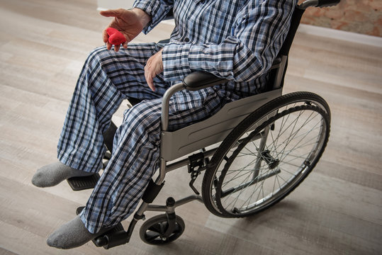 Old man holding a red small toy in the shape of heart. He is wearing pajamas and sitting in wheelchair in room. Close up