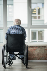 Senior male is sitting in wheelchair in front of the window in room. He is looking outside. Concept...