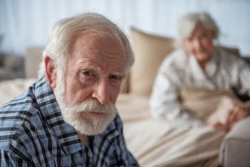 Portrait of depressed old pensioner looking at camera with upset look. Focus on his face. His wife...