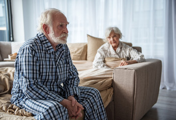 Upset old man sitting on bed with sad look. His woman sitting in bed under the blanket and looking...