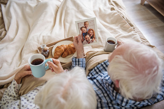 Top view of old husband and wife sitting in bed. They are holding cups of tea and photo in frame