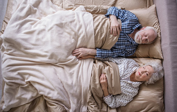 Top view of serene married couple having nap at home. Mature woman and man lying in bed and holding hands of each other