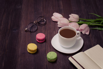 Obraz na płótnie Canvas Cup of tea, macarons, glasses, pink tulips and notebook on wooden background.