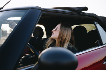 Fototapeta na wymiar Beautiful and hip young blonde woman or teenager, hipster or millennial sits inside convertible car and opens electronical button roof top of cabriolet. First vehicle and driving license practice