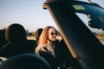 Fototapeta na wymiar Attractive, beautiful and sexy young blonde woman sits inside convertible cabriolet car, fixes hair and looks into rearview mirror during sunset. Wears stylish and fashionable vintage sunglasses