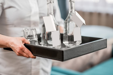 Chanmbermaid holding a tray with water in the glass bottles in the hotel bedroom. Close-up view without face
