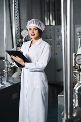 beautiful young girl in white working clothes is making a note on paper against the background of factory equipment