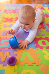 Cute baby in white vest, reaching for cups while on lying on tummy on alphabet mat.