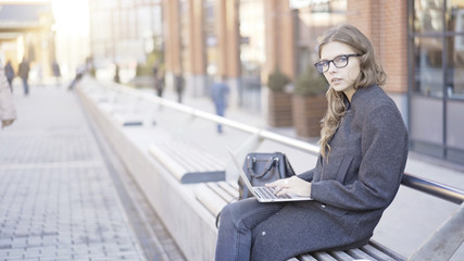 Beautiful young woman sitting with her laptop on a street bench and looking at the viewer