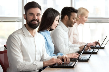 Obraz premium Cheerful smiling bearded caucasian man in office. Young adult bearded man smiling sitting in bright office woth colleagues.