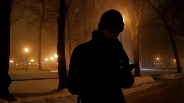 4k Mobster look dramatical man with black coat reads messages by mobile phone in winter night park