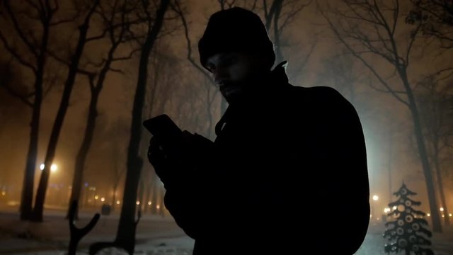 4k Mobster look man with black coat reads messages by mobile phone in winter cold night park close-up