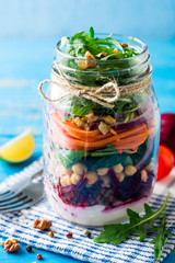 Healthy homemade salad with chickpeas, vegetables and walnuts in glass jar on blue background.