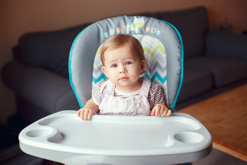 Portrait of cute adorable blonde Caucasian child girl sitting in high chair ready to eat. Everyday lifestyle. Candid real authentic moment. Kid one year old looking in camera