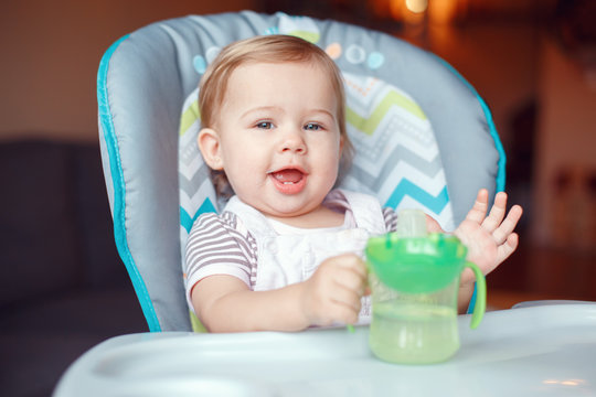 Portrait of cute adorable Caucasian child kid girl sitting in high chair drinking water from spill-proof sippy cup. Everyday lifestyle. Candid real authentic moment