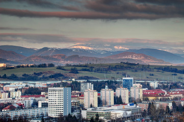 Obraz premium Row of socialist era tower blocks and houses in Banska Bystrica outskirts with snow-capped sunlit Velka Chochula peak of Nizke Tatry mountain range at winter sunset from Urpin hill Slovakia Europe