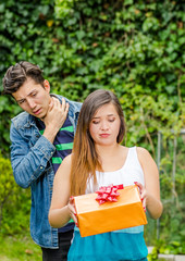 Close up of young woman doing a face of detesting the gift she is holding in her hands, with thoughtful boyfriend behind with worried face, friend zone concept