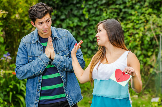 Close up of young woman with a disgusting face holding in her hands a letter, with a worried boyfriend looking the girl and begging to be his girlfriend, friend zone concept