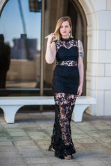 Blond alternative model gently twirling her hair while standing in black lace evening gown.