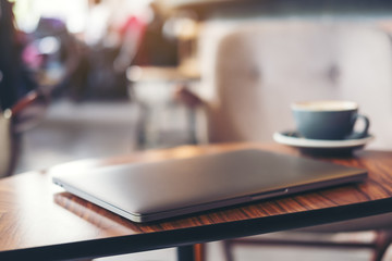 Closeup image of silver color laptop and coffee cup on wooden table with blur cafe background