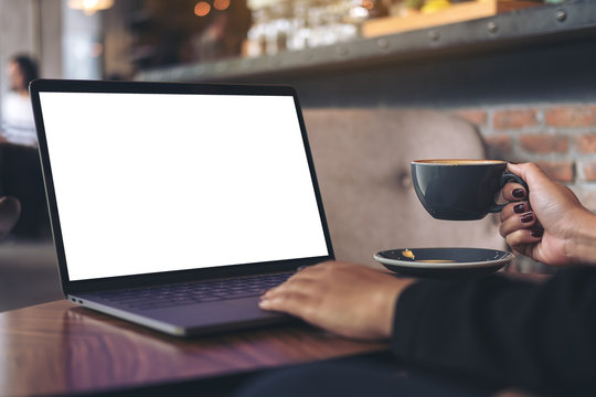 Mockup image of a businesswoman using laptop with blank white desktop screen while drinking hot coffee on wooden table in cafe