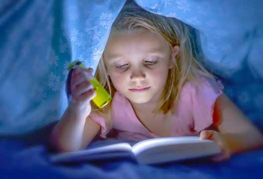 sweet beautiful and pretty little blond child girl 6 to 8 years old lying under bed covers reading book in the dark with torch flashlight smiling happy