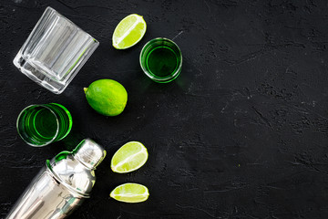Make cocktail with absinthe. Shaker, shots, lime slices on black background top view space for text