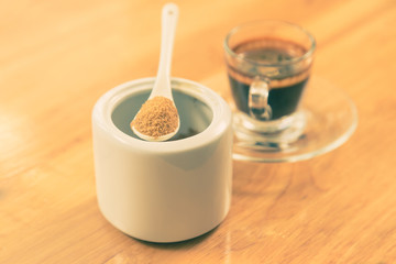 Brown sugar in a white ceramic spoon sitting on top of white ceramic container with blur coffee cup in background