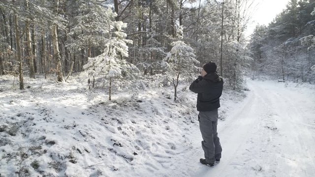 A young man is photographing a fur-tree in a winter forest.