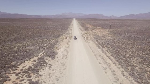 Aerial of SUV on straight dust road in desert driving towards mountains.