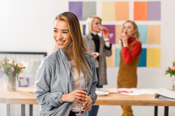 successful asian magazine editor with glass of champagne in modern office with coworkers behind