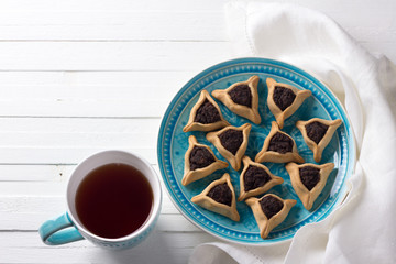 Traditional sweets for the Jewish holiday Purim. Hamantashen cookies or ears of Haman, triangular cookies with poppy seeds and raisins on a blue plate with a cup of tea, top view, free space