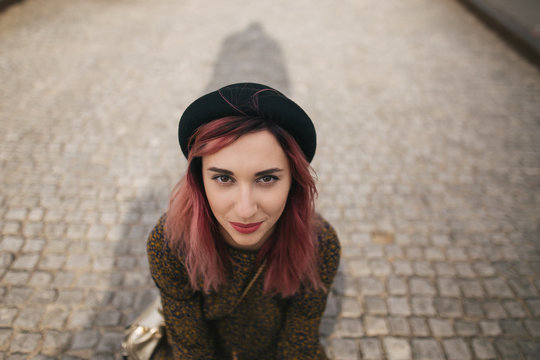 young girl with pink hair in trendy hat sitting on pavement in city