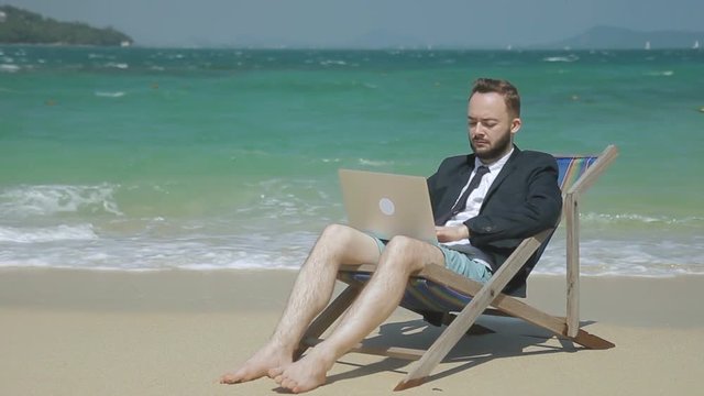 Young businessman is doing work using laptop while sitting on seashore. Bearded man is in work ing process, in deck chair on picturesque shore. Handsome guy in suit with tie has good time on beautiful