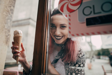 Happy young woman holding tasty ice cream in waffle cone and looking at camera through glass door...
