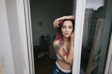 beautiful tender young woman in bra and denim shorts looking at camera through open window