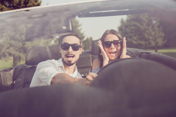 Oh no! Wtf! Front view, windscreen, guy grabs steering wheel while lady driver is panicking, both well dressed, in black trendy eyewear, woods around, sunny day