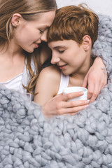 close-up shot of happy lesbian couple with cup of coffee embracing under knitted wool blanket