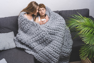 young lesbian couple sitting under knitted wool blanket on couch with cup of coffee
