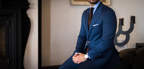Man in expensive tailored suit sitting on couch with his hands on his legs and posing in luxury apartment