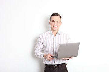 Smiling confident young man, no tie, holding grey laptop device and typing while standing against solid white wall. Wireless internet, wifi, enter password. Shaved, no facial hair. Isolated background