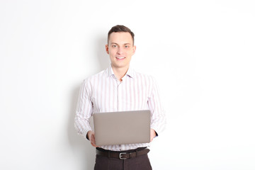 Smiling confident young man, no tie, holding grey laptop device and typing while standing against solid white wall. Wireless internet, wifi, enter password. Shaved, no facial hair. Isolated background