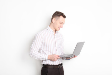 Smiling confident young man, no tie, holding grey laptop device and typing while standing against solid white wall. Wireless internet, wifi, enter password. Shaved, no facial hair. Isolated background
